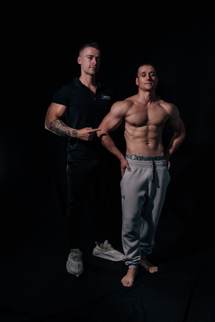 Two athletic men, one shirtless, posing in a dimly lit studio setting, showcasing muscular physiques.